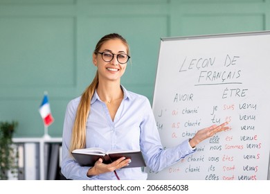 Happy French teacher explaining foreign language rules near blackboard indoors and smiling at camera. Confident young female tutor teaching linguistic online course