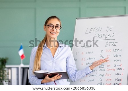 Happy French teacher explaining foreign language rules near blackboard indoors and smiling at camera. Confident young female tutor teaching linguistic online course