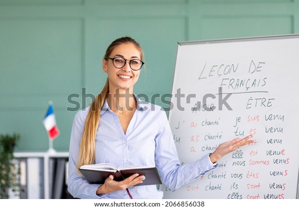 Happy French teacher\
explaining foreign language rules near blackboard indoors and\
smiling at camera. Confident young female tutor teaching linguistic\
online course