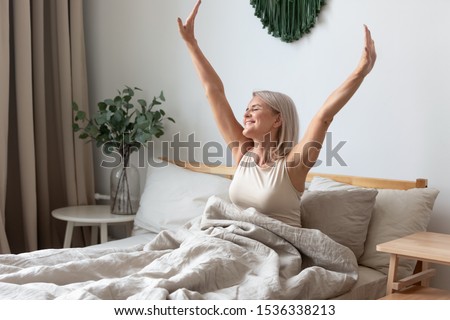 Happy fresh mature middle aged woman stretching in bed waking up alone happy concept, smiling old senior lady awake after healthy sleep sitting in cozy comfortable bedroom interior enjoy good morning