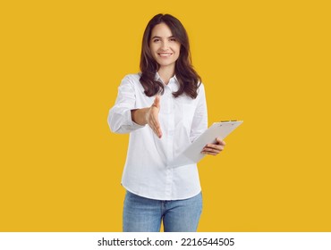 Happy friendly attractive young woman offering handshake. Pretty business lady in white shirt and jeans standing isolated on yellow background, holding clip board, extending her hand and smiling - Powered by Shutterstock