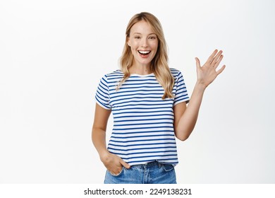 Happy and friendly blond girl smiling, waving hand and saying hello, hi gesture, greeting you with carefree relaxed face expression, white background - Powered by Shutterstock