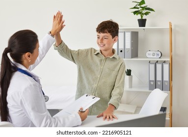 Happy Friendly Doctor And Child Patient Give Each Other High Five After Medical Checkup At Clinic. Little School Boy Smiling And High Fiving His Pediatrician While Standing By Her Desk In The Office