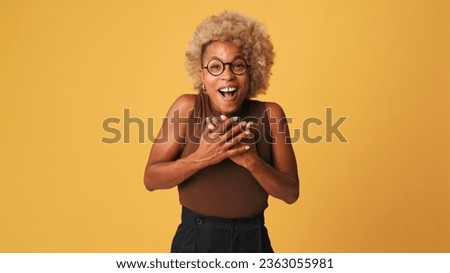 Happy friendly girl in glasses, wearing brown top looking at camera in pleasant joyful surprise, isolated on orange background, studio shot