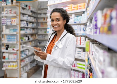 Happy Friendly Multiethnic Pharmacist Doing Inventory In A Provided And Modern Pharmacy While Looking At Camera. Portrait Of Smiling African Doctor Woman Working In Drugstore With Digital Tablet.