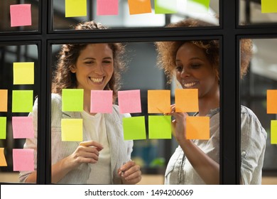 Happy friendly professional team two diverse businesswomen laugh talk check project progress on stickers, creative young female coworkers having fun planning project write on sticky notes glass wall