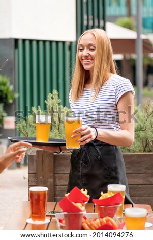 Happy friendly waitress serving pints of lager or beer at an outdoor restaurant smiling as the customer stretches out a hand to take the glass