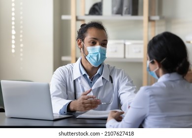 Happy Friendly Young African American Male Doctor Therapist Physician And Female Indian Patient In Facial Masks Respirators Communicating Sitting At Table In Clinic Office, Medical Healthcare Concept.