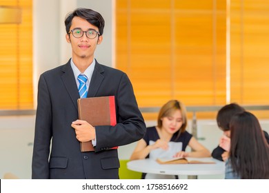 Happy friendly young asian teacher or pst graduate male student in glasses standing and holding book in front group of friend.