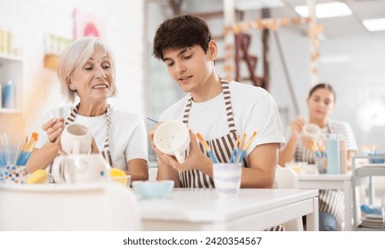 Happy friendly young guy sitting with older female colleague at table in pottery workshop, talking cheerfully while decorating ceramic mugs, applying design with paints.. - Powered by Shutterstock