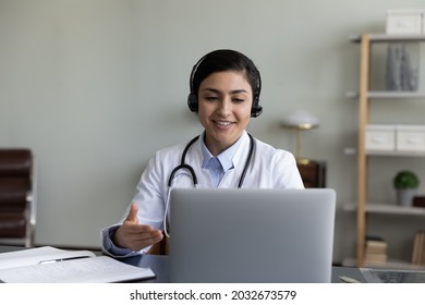 Happy friendly young indian gp doctor physician wearing headset with mic, giving distant healthcare consultation to patient on computer by video call or taking part in educational virtual event.