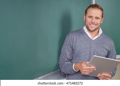 Happy friendly young teacher or pst graduate male student standing holding a tablet in front of a blank chalkboard