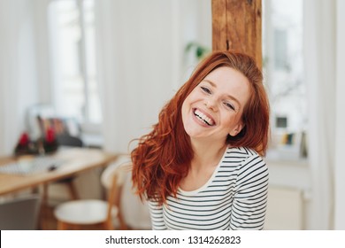 Happy friendly young woman with cute smile laughing with head to the side as she looks at camera indoors in her apartment