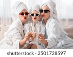 Happy friends in bathrobes with sunglasses and glasses of sparkling wine indoors, selective focus. Spa party