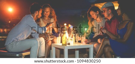 Happy friends celebrating at evening beach party outdoor drinking champagne - Young people having fun at  bar next to the ocean - Soft focus on center bottles - Youth lifestyle and summer concept