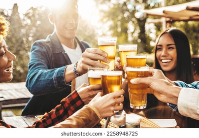 Happy friends cheering beer glasses at brewery pub garden - Group of young people enjoying happy hour sitting at bar table - Life style concept with guys and girls having dinner party together - Powered by Shutterstock