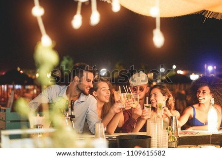 Happy friends cheering and drinking cocktails at beach party outdoor - Young millennials people having fun at weekend summer night - Youth lifestyle and nightlife concept - Main focus on left guys
