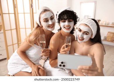 Happy friends with facial masks and glasses of sparkling wine taking selfie in bathroom. Spa party - Powered by Shutterstock