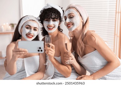 Happy friends with facial masks and glasses of sparkling wine taking selfie in bathroom. Spa party - Powered by Shutterstock