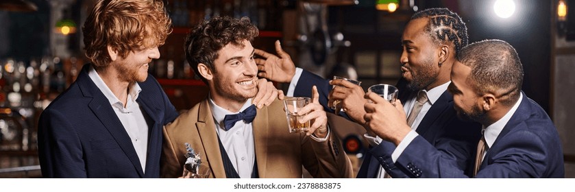 happy friends in formal wear congratulating groom in bar, interracial men holding whiskey banner