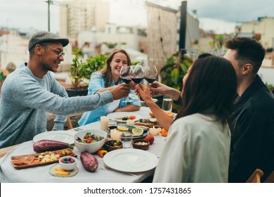 Happy Friends Having Dinner Party Outside Sitting In Cozy Restaurant Terrace With A Garden Celebrating A Long-awaited Meeting Or Birthday Making Toasts With Red Wine, Laughing And Having Fun Together