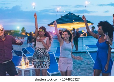 Happy friends having fun beach party outdoor with fireworks - Young people drinking champagne and dancing together in summer festival - Soft focus on right woman face - Youth and summer concept
