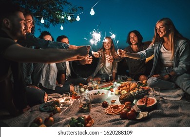 Happy Friends Having Fun With Fire Sparkles. Young People Camping At Picnic On The Night. Youth Friendship Concept On Night Mood