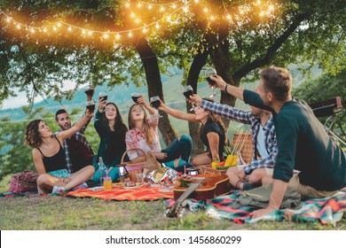 Happy Friends Having Fun At Picnic Dinner With Vintage Lights Outdoor Next Vineyard - Young People Cheering With Red Wine On Weekend Summer Night - Friendship, Party Concept - Focus On Left Guys Faces