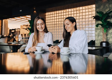 Happy friends having fun together on a coffee break in a cafe. A young woman is holding a phone and showing pictures to her best friend. Two girls drinking coffee and having fun together. - Powered by Shutterstock