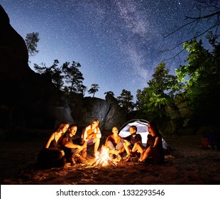 Happy Friends hikers having a rest together around campfire beside camp and illuminated tourist tent at night. On background beautiful night starry sky full of stars and Milky way, mountain rocks. - Powered by Shutterstock