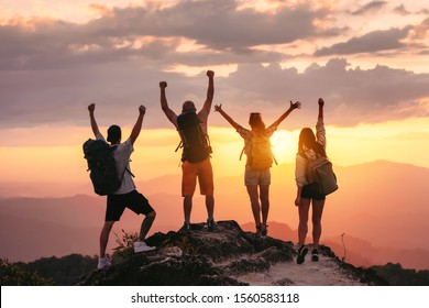 Happy friends hikers or tourists stands with raised arms on mountain top against mountains and looking at sunset