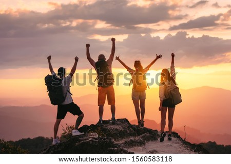 Happy friends hikers or tourists stands with raised arms on mountain top against mountains and looking at sunset