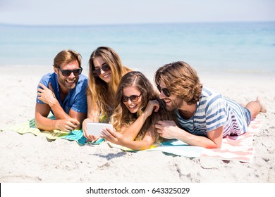 Happy Friends Looking In Mobile Phone While Lying On Sand At Beach During Sunny Day