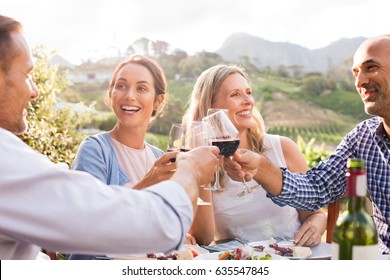 Happy Friends Raising Their Glasses In A Toast  In A Winery Farm. Smiling Mature Woman And Men Enjoying A Picnic Together. Middle Aged Multiethnic Couple Having Dinner Together And Toasting Wine.