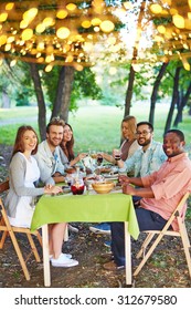 Happy Friends Sitting By Thanksgiving Table And Looking At Camera Outdoors