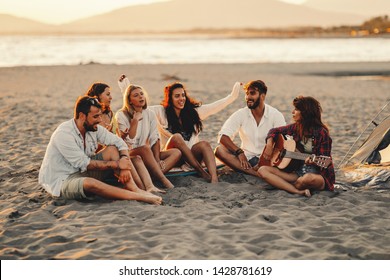 Happy friends sitting on the beach singing and playing guitar during the sunset - Powered by Shutterstock