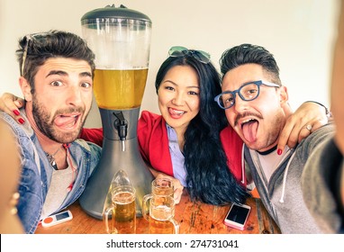 Happy friends taking selfie with funny tongue out drinking beer from tower dispenser - Friendship fun concept with new trends and technology -  Alternative everyday party life in vintage brewery bar