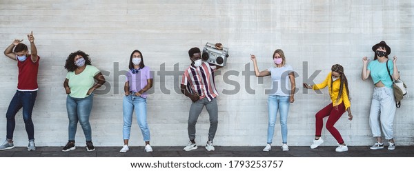 Happy friends wearing face mask listening music\
with vintage boombox outdoor - Multiracial young people having fun\
dancing together during corona virus outbreak - Youth millennial\
friendship concept