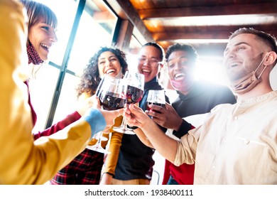 Happy friends wearing protective face masks toasting red wine sitting at bar restaurant - New normal friendship concept with young people having fun at home party - Focus on glasses