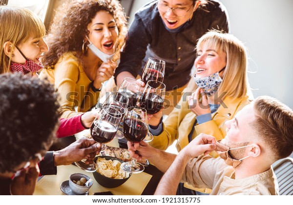 Happy friends\
wearing protective face masks toasting red wine sitting at\
restaurant - New normal friendship concept with young people having\
fun at home party - Focus on\
glasses