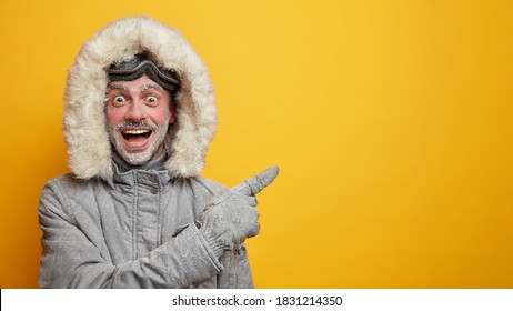 Happy Frozen Man In Warm Clothes Has Fun During Winter Points Away On Empty Space Wears Gloves Isolated On Yellow Background. Cheerful Male Adventurer With Frosted Beard And Ski Goggles Has Expedition