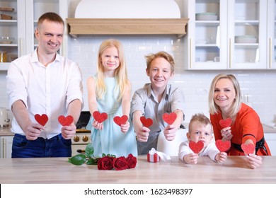 Happy Full Family Of Dad Mom And Three Kids Two Boys And Girl Is Holding Red Paper Hearts And Smiling, Family Is Standing In Kitchen At Home, Blonde Caucasian People. Valentines Day And Love