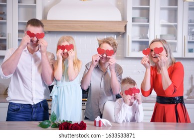 Happy Full Family Of Dad Mom And Three Kids Two Boys And Girl Is Holding Red Paper Hearts And Smiling, Family Is Standing In Kitchen At Home, Blonde Caucasian People. Valentines Day And Love