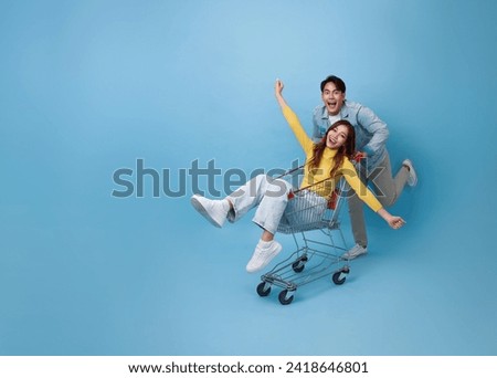 Happy fun Asian woman sitting inside of shopping trolley and man pushing shopping cart to get the latest offers promotion at the supermarket isolated on blue background.