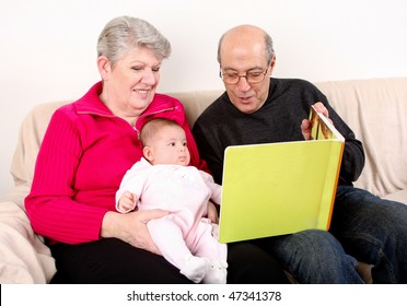 Happy Fun Caucasian Hispanic Middle Eastern Family Sitting Together On Couch Reading Book To Baby. Grandfather And Grandmother Reading Green Book To Infant Granddaughter.