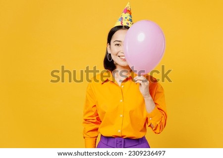 Happy fun cheerful smiling young woman wears casual clothes hat celebrating covering half of face with pink balloon look camera isolated on plain yellow background. Birthday 8 14 holiday party concept