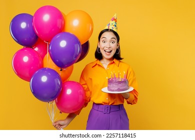 Happy fun excited amazed young woman wears casual clothes hat celebrating hold bunch of colorful air balloons cake with candles isolated on plain yellow background. Birthday 8 14 holiday party concept