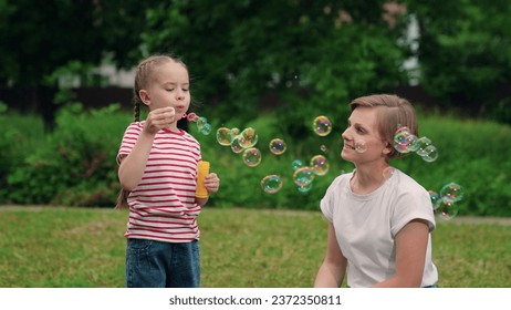 Happy fun kid, girl, mother blows soap bubbles against background of green trees. Daughter, mom play with soap bubbles in park summer day. Happy family relaxing in nature. Concept of child's play. - Powered by Shutterstock