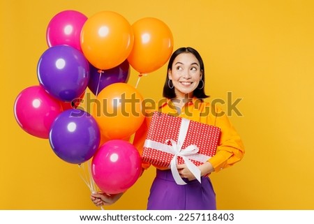Happy fun smiling amazed young woman wear casual clothes celebrating look at balloons holding present box with gift ribbon bow isolated on plain yellow background. Birthday 8 14 holiday party concept