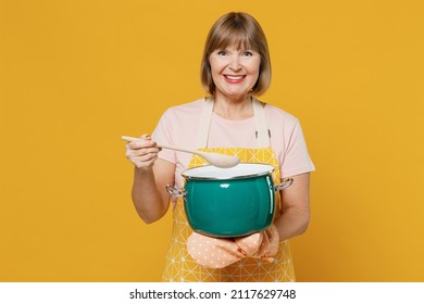 Happy Fun Smiling Satisfied Elderly Housekeeper Housewife Woman 50s In Orange Apron Hold Spoon Green Saucepan With Soup Isolated Plain On Yellow Background Studio. People Household Lifestyle Concept